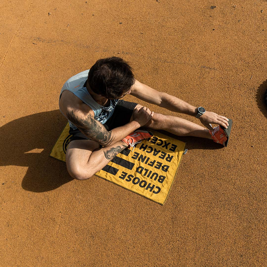 Homem a fazer alongamentos sentado ao ar livre sobre uma toalha da GoldNutrition com as palavras 'Choose, Build, Define, Reach, Excelence', simbolizando a importância da recuperação ativa após treinos ou provas de alta intensidade.