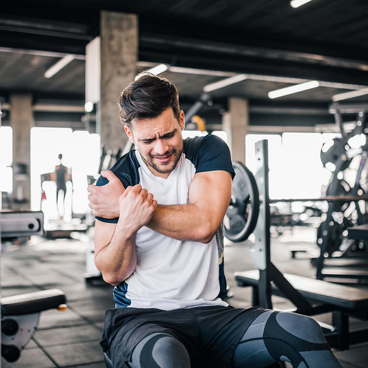 Homem jovem com expressão de dor segurando o braço no ginásio. Ele veste uma t-shirt branca e preta e calções pretos, sentado num banco de musculação com equipamentos de treino ao fundo.